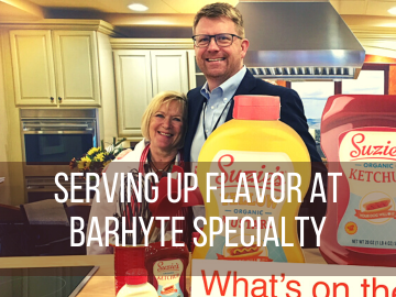 A tall man with glasses stands with his arm around his mother in a kitchen behind a cardboard display of ketchup and mustard bottles. The super-imposed text reads "Serving Up Flavor at Barhyte Specialty"