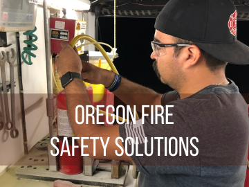 A man with safety glasses and a backwards baseball cap attaches a hose to the top of a fire extinguisher on a workbench.